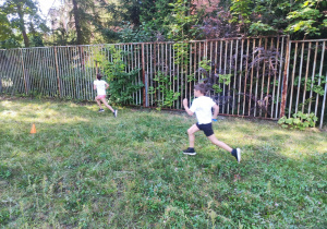 Dwóch uczniów rywalizuje na trasie biegu. W tle ogrodzenie. Trasa ograniczona pachołkami.